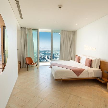 Five Jumeirah Village Dubai Exterior photo The photo shows a modern bedroom with a minimalist design. There is a large bed with white bedding and pink accents, positioned against a wall. To the side, there is a round mirror and a stylish chair. The room features large windows dressed with lig