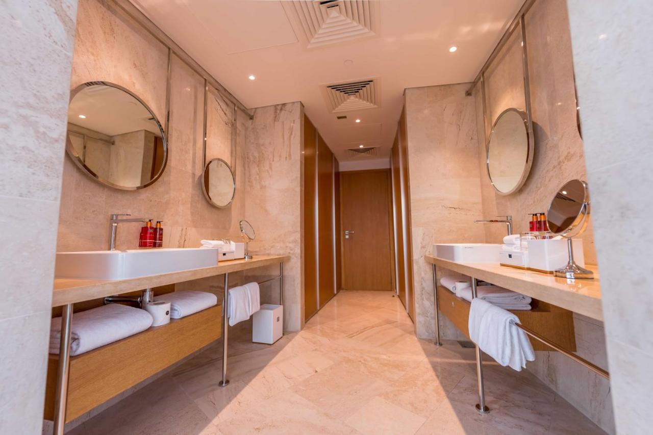 Five Jumeirah Village Dubai Exterior photo The photo shows a modern bathroom featuring dual vanities. Each vanity is equipped with a white sink and has a circular mirror above it. The countertops are made of a light-colored stone, and there are wooden cabinets underneath that hold rolled whit