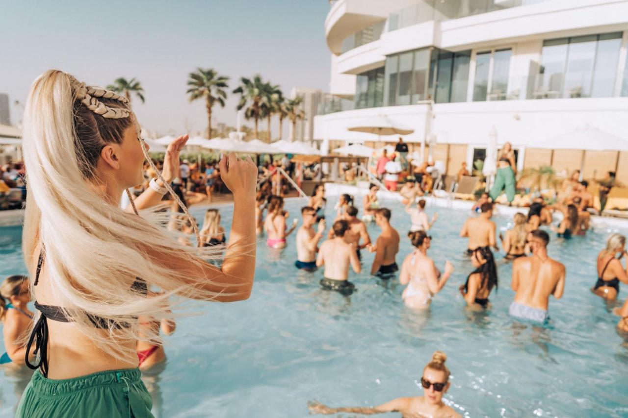 Five Jumeirah Village Dubai Exterior photo The photo features a lively pool party scene. In the foreground, a woman with long, blonde hair is seen raising her arm as if enjoying the moment. The pool is filled with many people, some swimming and others socializing. The atmosphere appears festi