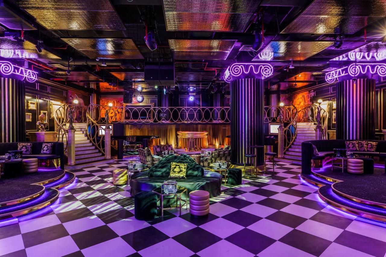 Five Jumeirah Village Dubai Exterior photo The photo depicts a stylish, upscale nightlife venue, likely a nightclub or lounge. The interior features a checkered floor in black and white, creating a striking visual effect. There are plush seating arrangements with green and purple accents, sug