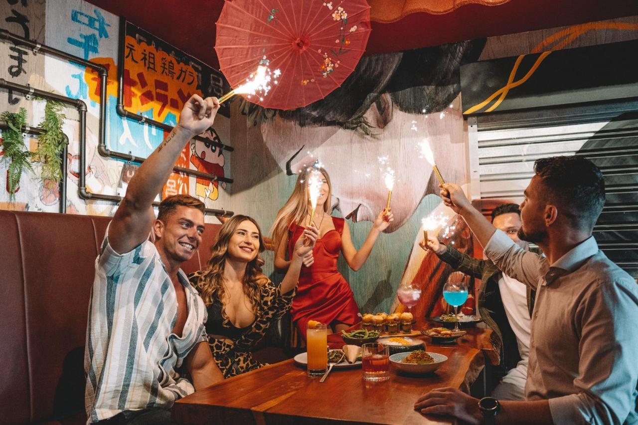 Five Jumeirah Village Dubai Exterior photo The photo depicts a lively gathering at a dining table where several people are celebrating. There are four individuals: two men and two women. One of the women is wearing a red dress, while the other is dressed in a patterned outfit. They are holdin