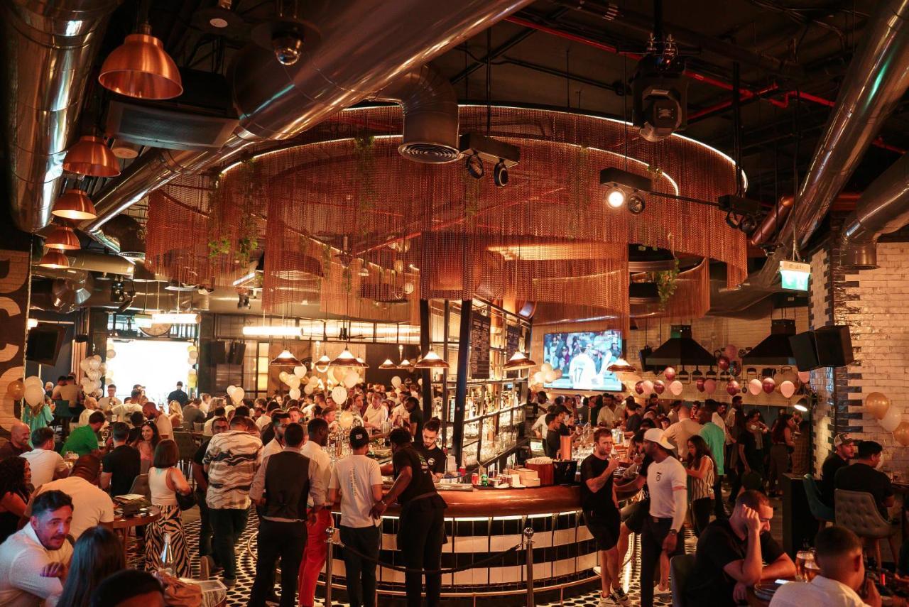 Five Jumeirah Village Dubai Exterior photo The photo depicts a lively bar or restaurant scene filled with people socializing. The interior design features a modern aesthetic with a mix of industrial elements, such as exposed pipes and metallic accents. A large, circular bar is the focal point