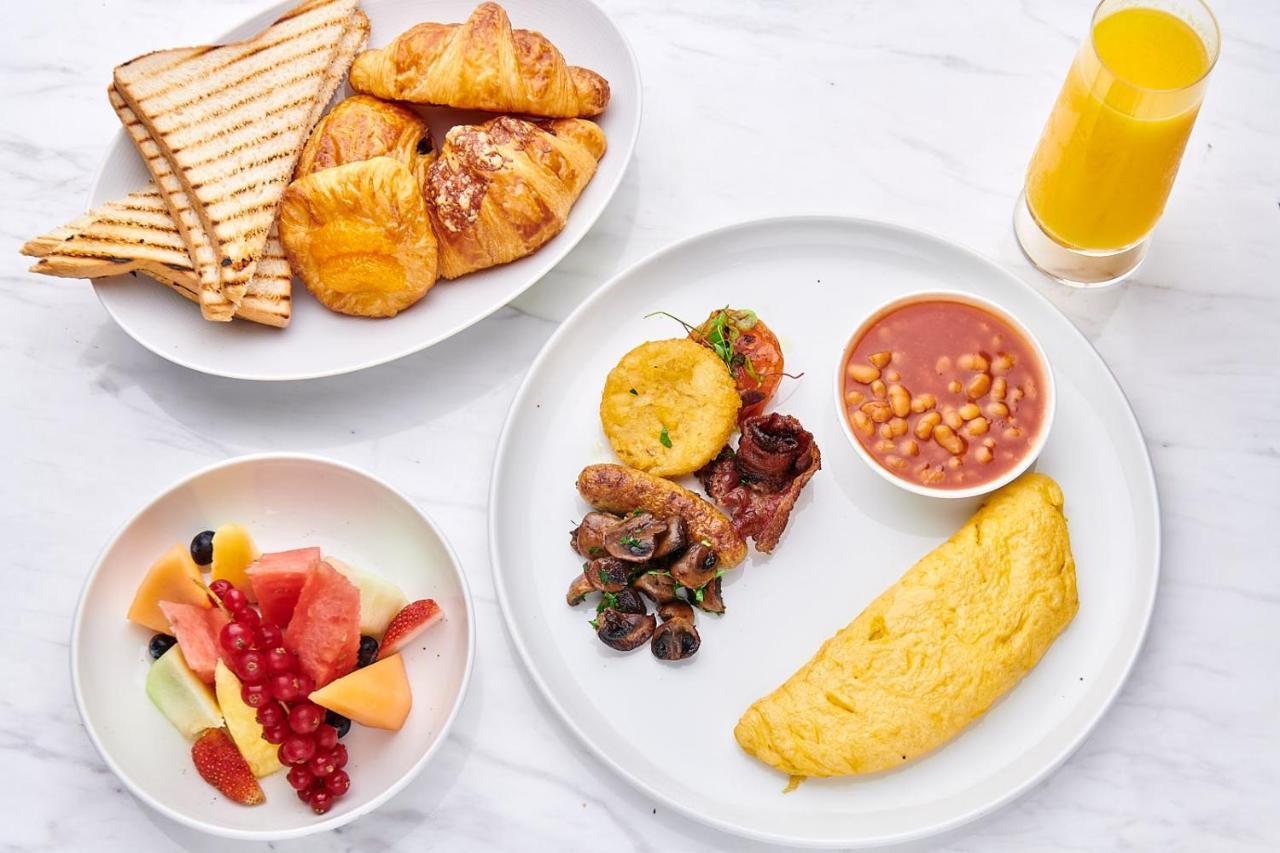 Five Jumeirah Village Dubai Exterior photo The photo displays a colorful breakfast spread. On a white plate, there is a fluffy omelet, a serving of baked beans, a yellow plantain slice, and a portion of sautéed vegetables, including tomatoes. Next to it, there is a smaller bowl with a variety