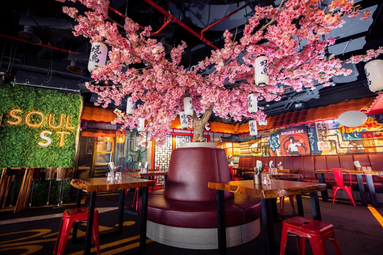 Five Jumeirah Village Dubai Exterior photo The photo shows a vibrant and modern restaurant interior. At the center, there is a large cherry blossom tree adorned with pink flowers and hanging lanterns, creating a lively atmosphere. Surrounding the tree is a circular booth with maroon upholster