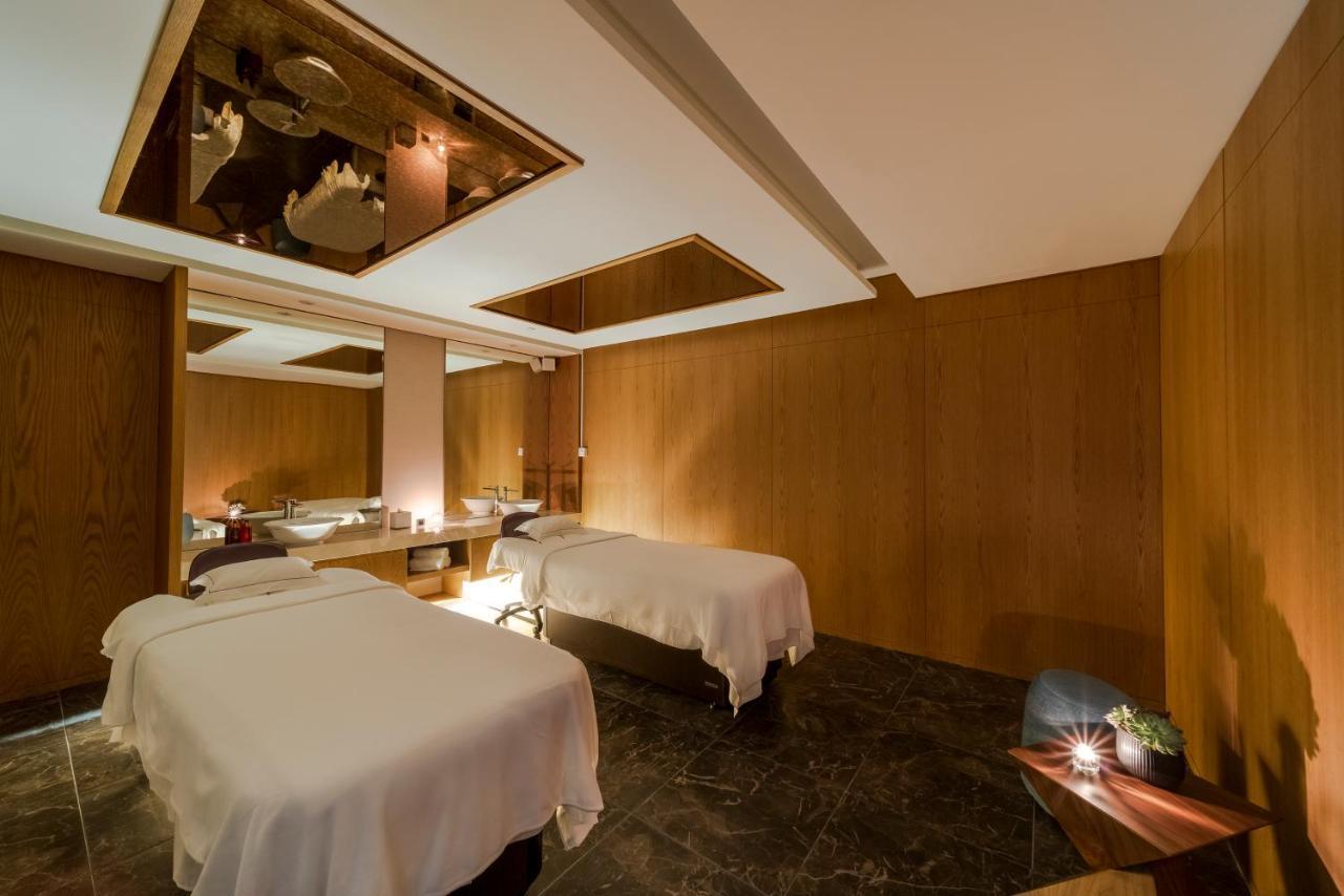 Five Jumeirah Village Dubai Exterior photo The photo shows a tranquil spa room designed for relaxation and wellness. There are two massage tables, each covered with white linens, positioned in the center of the room. The walls are finished with warm wood paneling, creating a cozy atmosphere. 