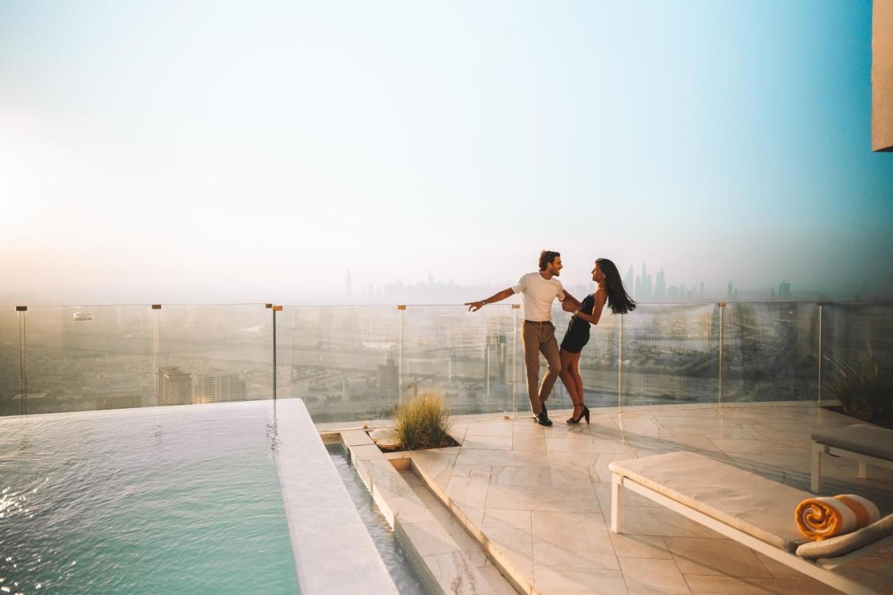 Five Jumeirah Village Dubai Exterior photo The image shows a couple dancing together on a terrace or balcony with a view of a city skyline in the background. They appear to be enjoying a romantic moment, surrounded by a luxurious setting that includes a swimming pool. The atmosphere is bright