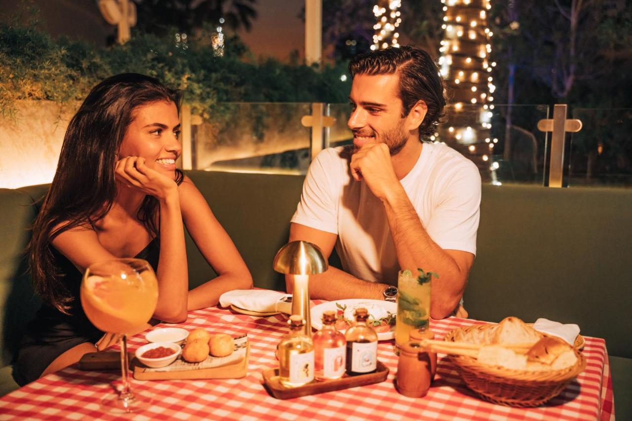 Five Jumeirah Village Dubai Exterior photo The photo shows a romantic dining scene featuring a couple at a table. The setting is illuminated with soft lights, creating a warm atmosphere. The table is adorned with a red and white checkered tablecloth, and various dishes are presented, includin