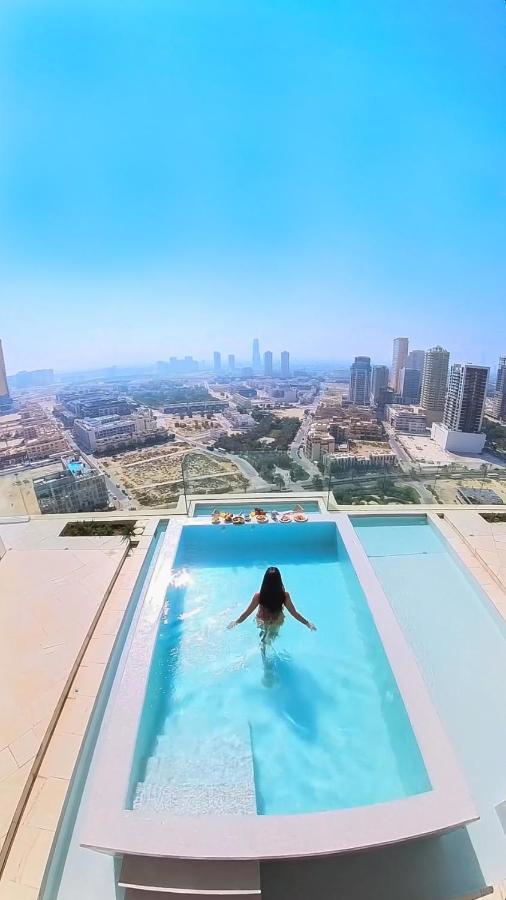 Five Jumeirah Village Dubai Exterior photo The photo depicts a person sitting in a swimming pool on a rooftop. The pool is positioned at the edge, offering a stunning view of a city skyline that includes tall buildings and a horizon stretching into the distance. The sky is clear and blue, sug