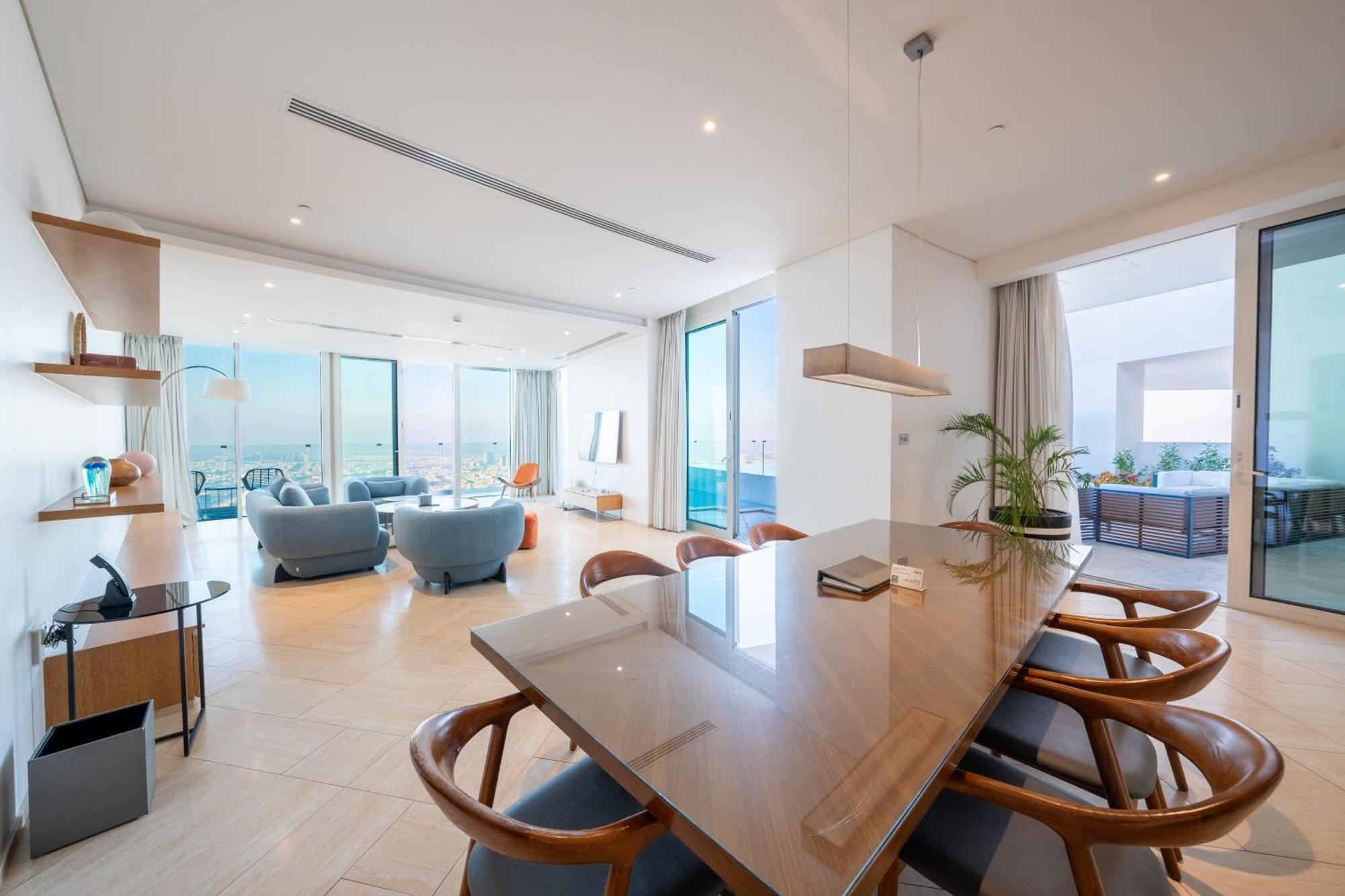 Five Jumeirah Village Dubai Exterior photo The photo displays a modern and stylish living space. In the foreground, there is a large wooden dining table with several wooden chairs around it. The dining area transitions into a spacious living room featuring large windows that offer a view of t