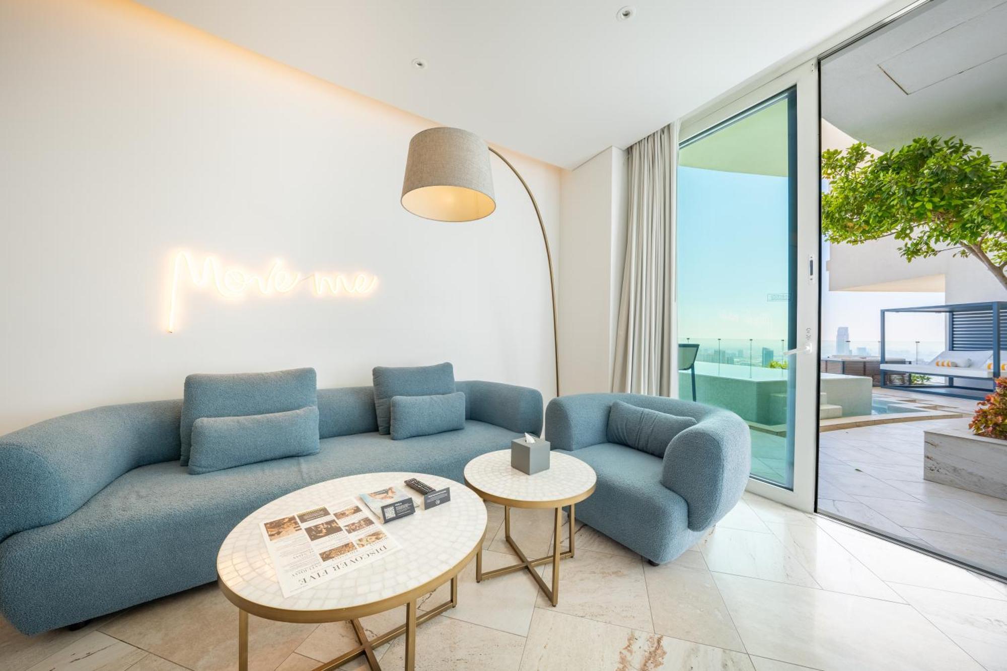 Five Jumeirah Village Dubai Exterior photo The photo shows a modern and stylish living space. It features a light blue sectional sofa and a matching armchair, creating a cozy seating area. Two round marble coffee tables are placed in front, one of which has some magazines or books on it. A ta