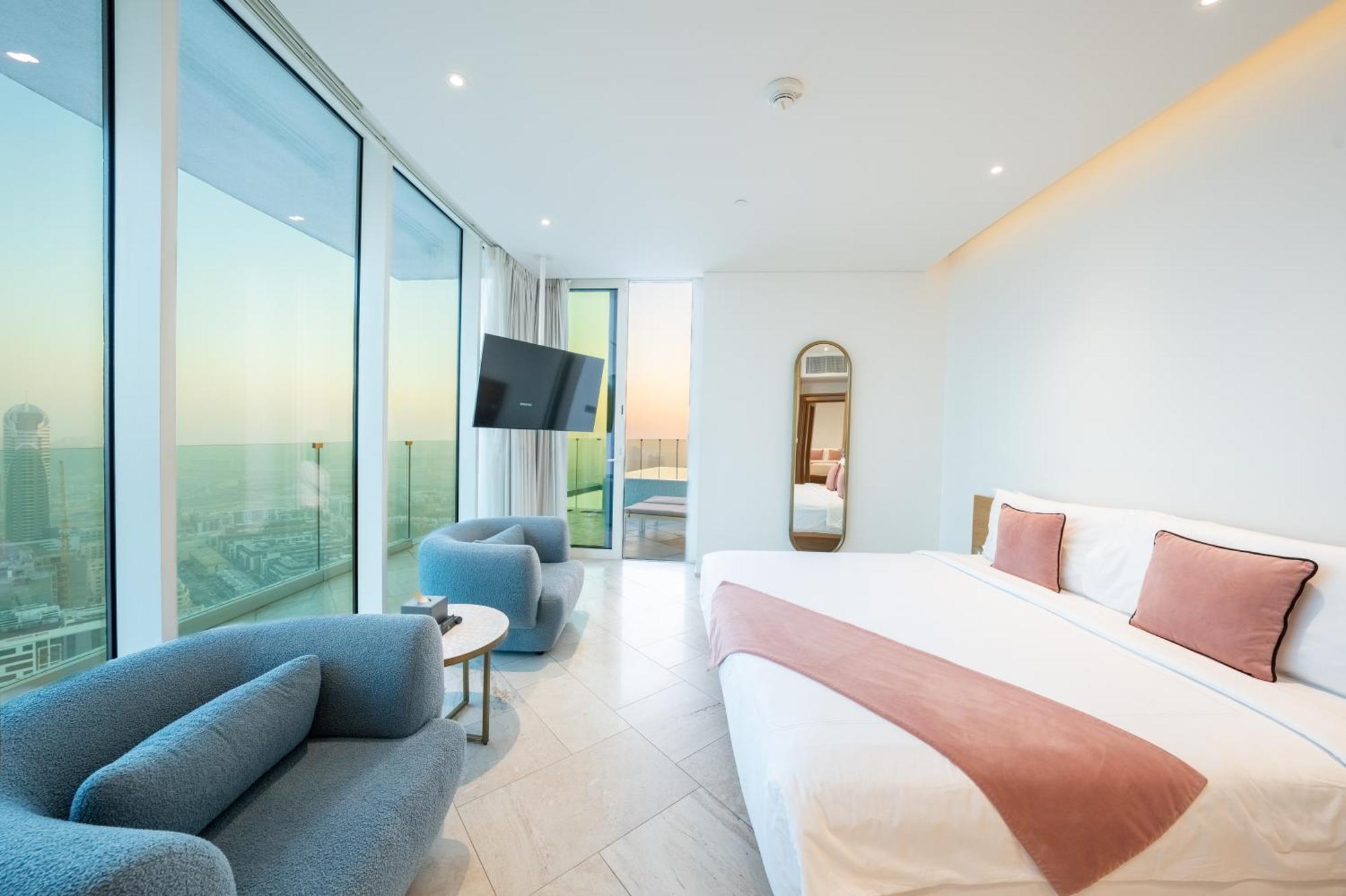 Five Jumeirah Village Dubai Exterior photo The photo depicts a modern hotel room featuring large windows that provide a view of the outside. The room includes a spacious bed with a pink accent blanket, a cozy blue sofa, a small round table, and a wall-mounted television. There is also a mirro