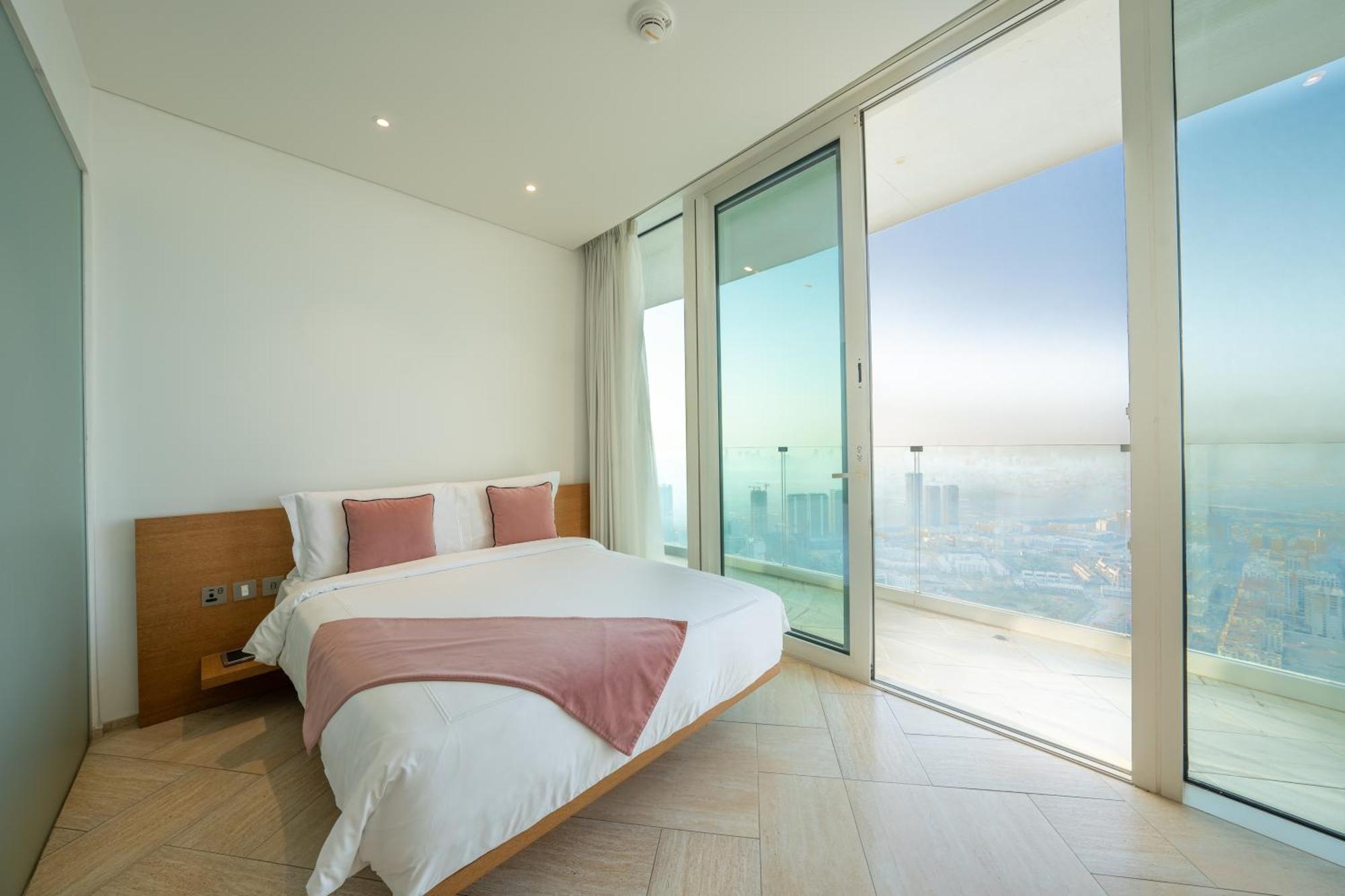 Five Jumeirah Village Dubai Exterior photo The photo shows a modern bedroom with a large bed featuring white bedding and accent pillows in a soft pink color. The room has floor-to-ceiling glass doors that lead out to a balcony, providing a panoramic view of the outside. The walls are painted 