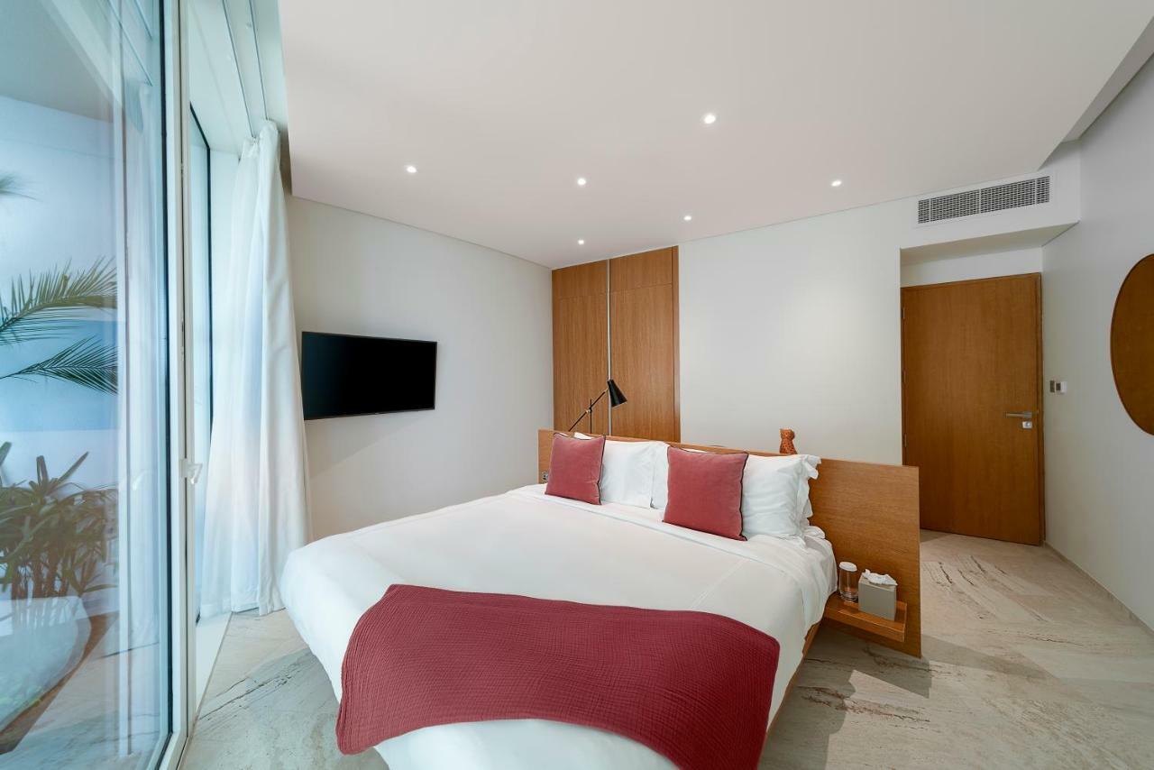 Five Jumeirah Village Dubai Exterior photo The photo shows a modern and stylish bedroom. It features a bed with white bedding and two red decorative pillows. There is a mounted flat-screen TV on the wall opposite the bed. The room has large glass windows that allow natural light to enter, and