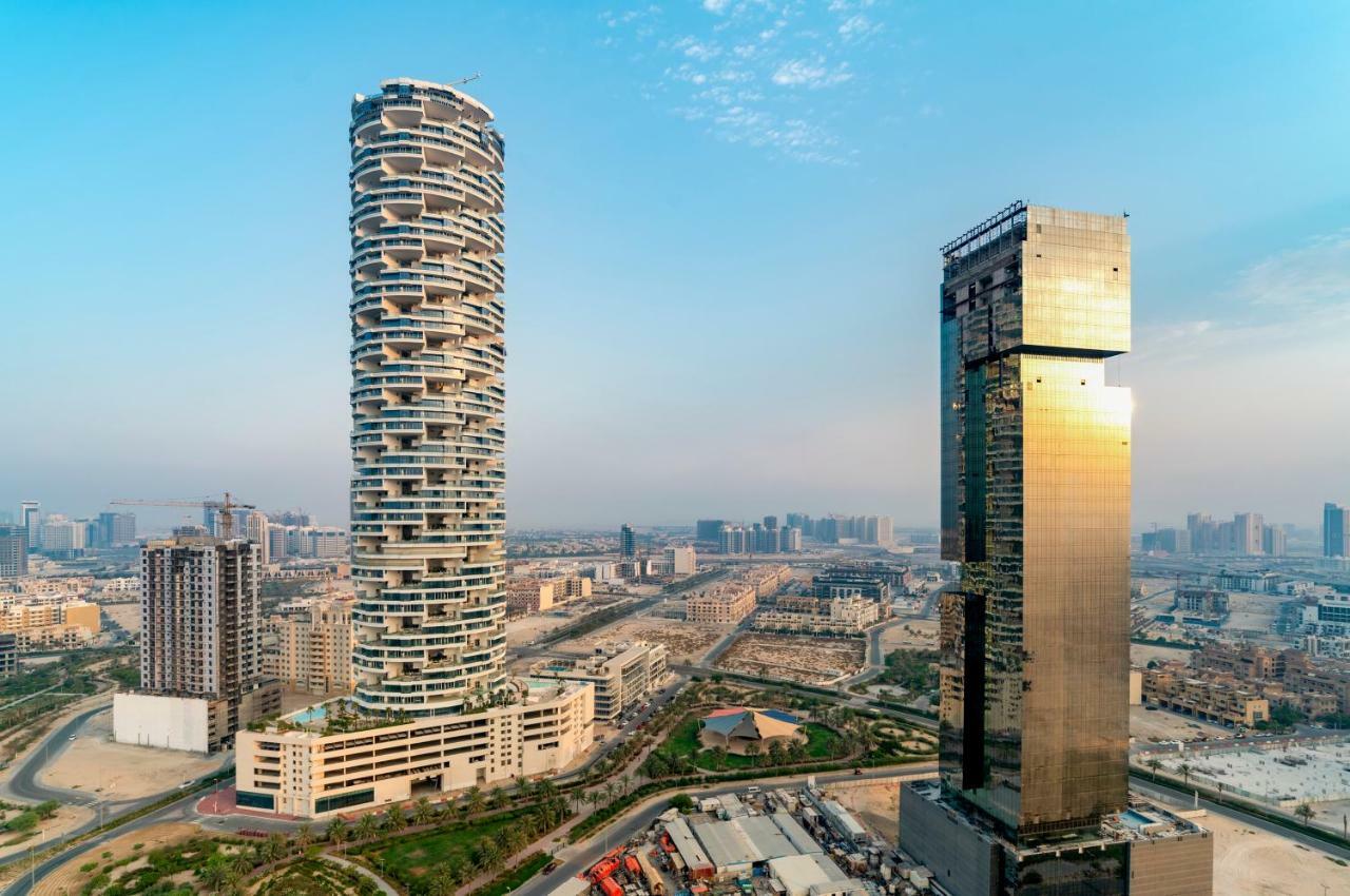 Five Jumeirah Village Dubai Exterior photo The photo shows two distinctive skyscrapers located in an urban area. On the left, there is a tall, cylindrical building with a unique, modern design characterized by its twisting, spiral shape and balconies at various levels. To the right, there is 