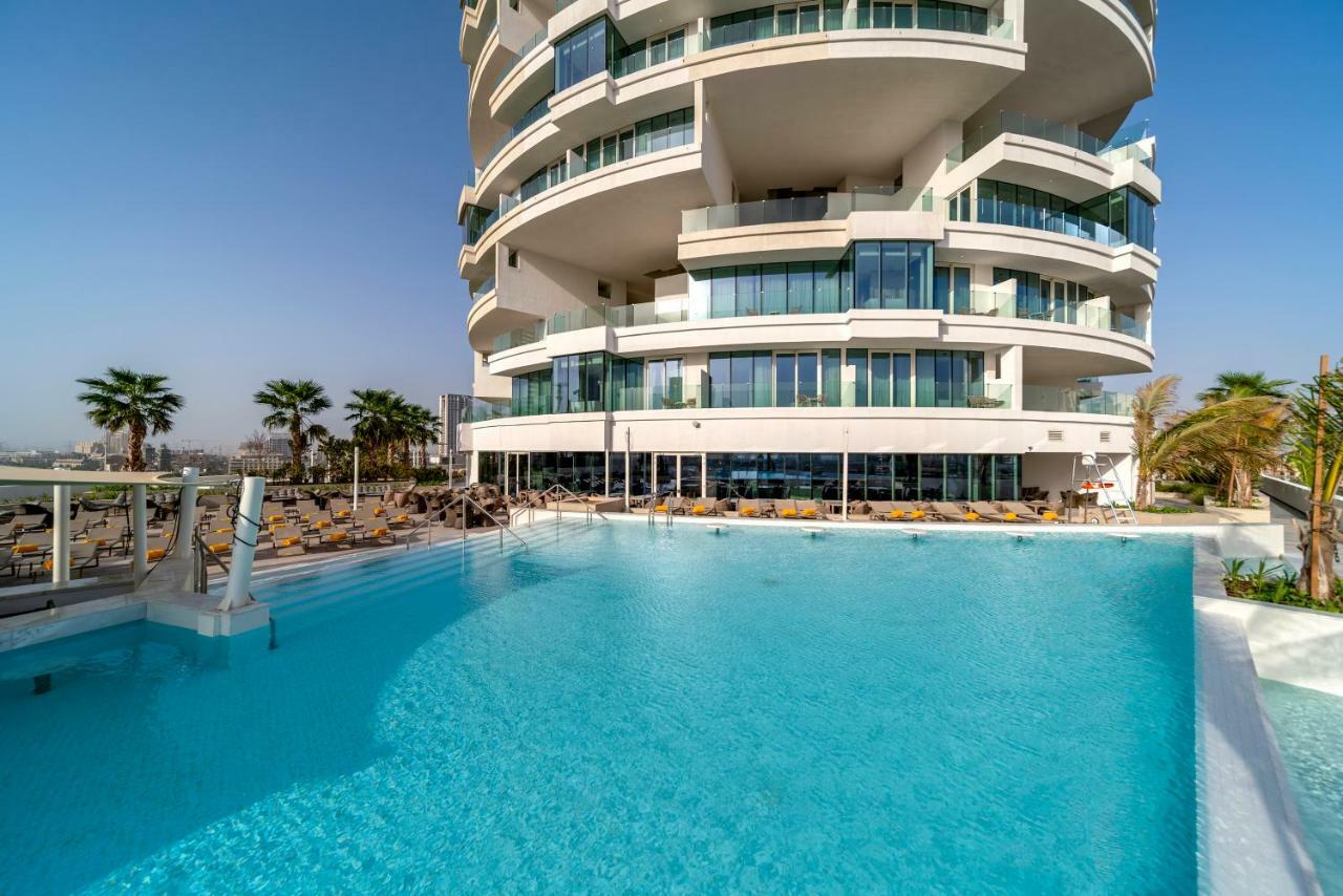 Five Jumeirah Village Dubai Exterior photo The photo depicts a modern building with a distinctive architectural design, featuring multiple curved balconies. In front of the building, there is a large, inviting swimming pool with clear, turquoise water. Surrounding the pool are loungers for re